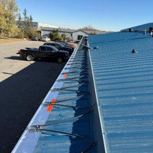 Commercial metal roof de-icing cable system installation with heavy-duty mounting clips on a blue industrial roof.