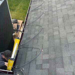 Professional contractor on a ladder installing eavestrough heating cables on an asphalt shingle roof profile.